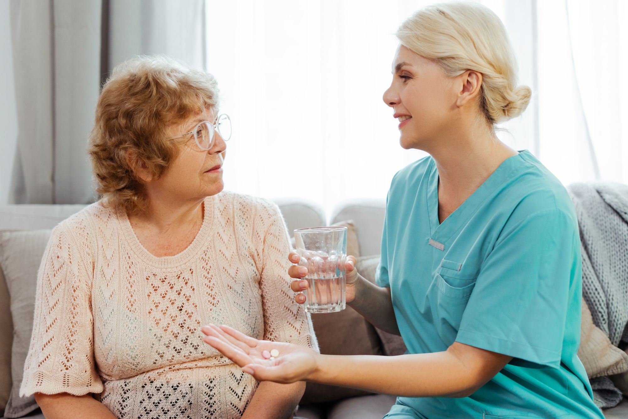 Nurse giving medication to a senior woman on the sofa holding glass with water