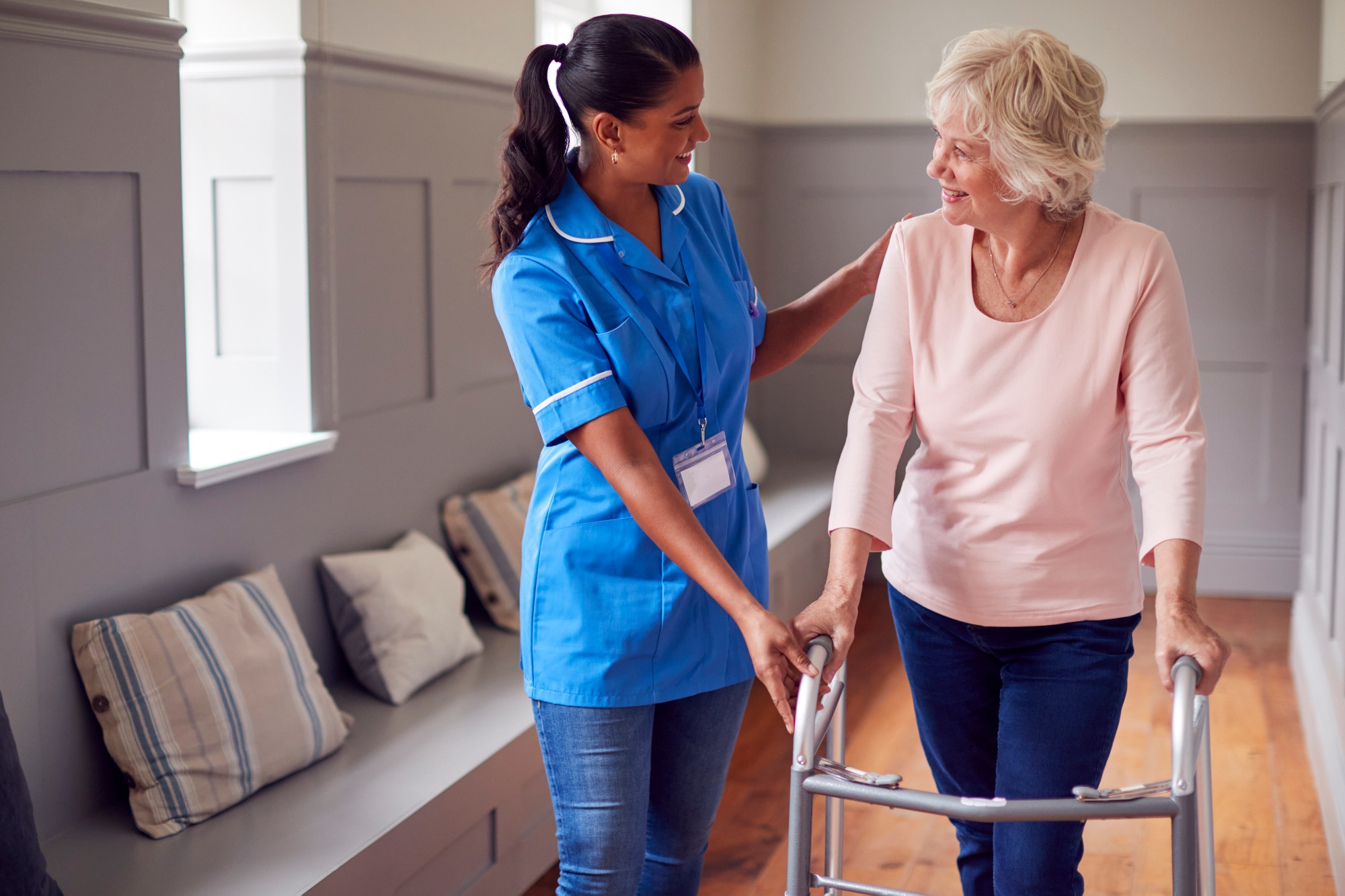 Senior Woman At Home Using Walking Frame Being Helped By Female Care Worker In Uniform
